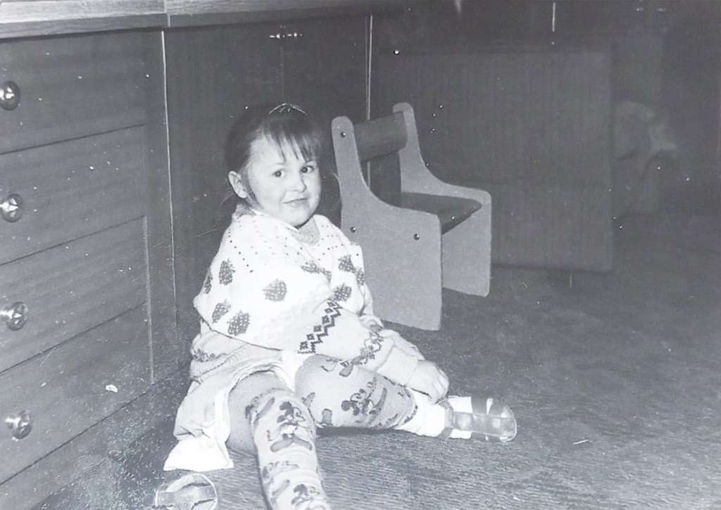 Me as a child. I'm sitting in our living room, trying to put my sandal on while looking straight to the camera. I'm wearing my favourite tights with Mickey Mouse and a sweater with strawberries. I was probably around six years old.