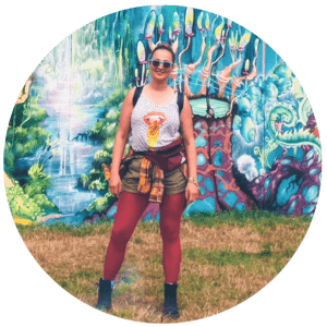 a girl in dark red tights, shorts and boots. She is wearing sunglasses and a vest. She's at a festival. Graffiti on the wall in the background.