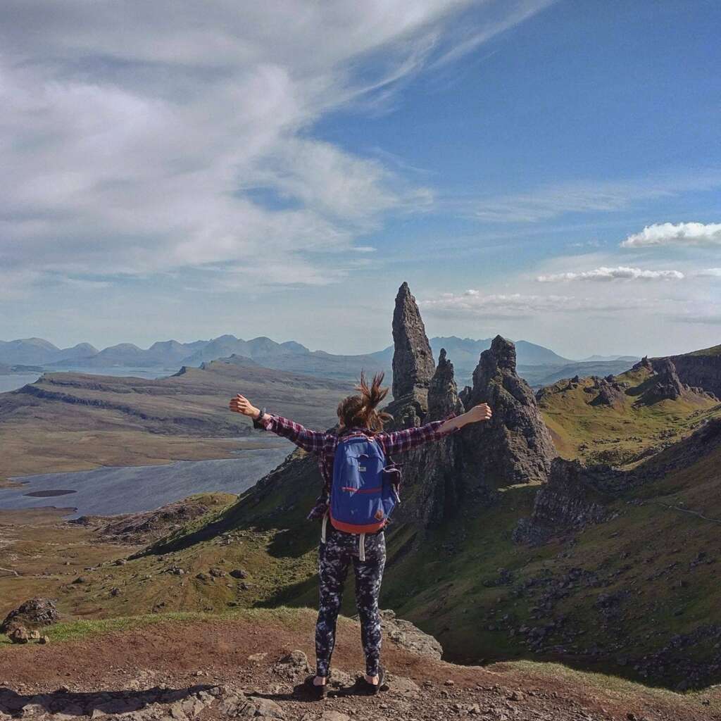 A woman standing on top of the Old Man of Storr in the Isle of Skye, Scotland, with arms raised high in celebration. The majestic landscape of the Scottish Highlands and the rocky peak of the Old Man of Storr can be seen in the background.