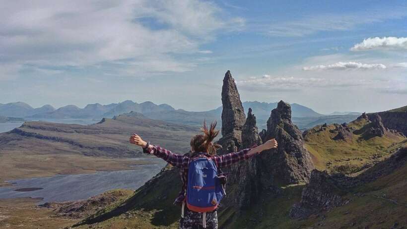 A woman standing on top of the Old Man of Storr in the Isle of Skye, Scotland, with arms raised high in celebration. The majestic landscape of the Scottish Highlands and the rocky peak of the Old Man of Storr can be seen in the background.